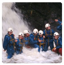 image of a larger group of children wearing buoyancy aids, helmets and waterproofs at the base of a waterfall looking like they are having alot of fun