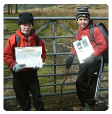  image of two boys stood in front of a gate wearing woolly hats, gloves and rucksacks facing the camera with their maps