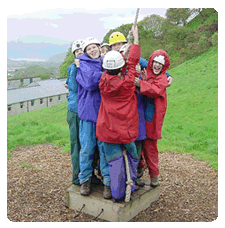 image of a group of children on a small wooden square platform holding on tight to each otehr having each swinging onto the platform using a rope swing