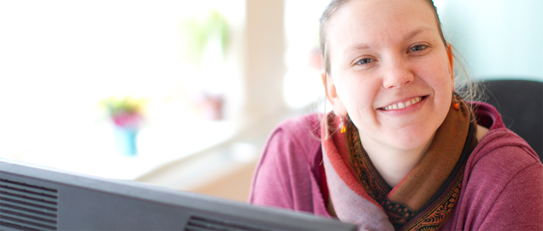 A photo of an adult sitting behind a monitor smiling at the camera