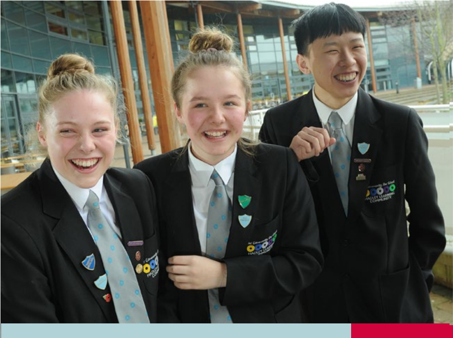 Three students in school uniforms standing outside a modern building with glass windows and wooden beams, wearing black blazers adorned with badges, white shirts, and light blue polka dot ties, engaged in conversation.