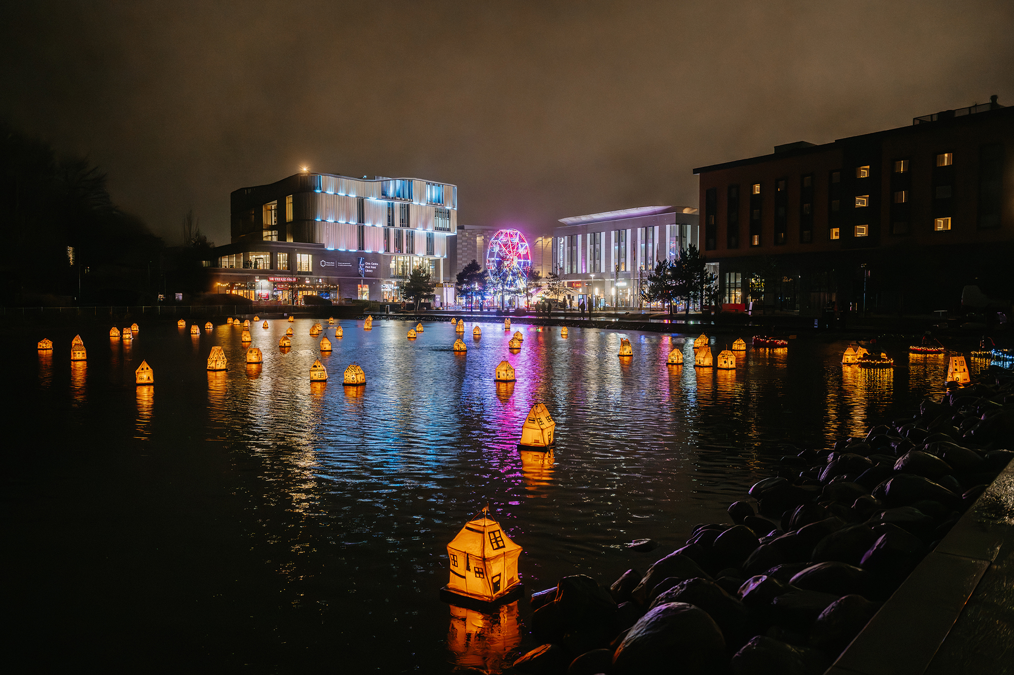 Dozens of glowing house-shaped lanterns float across a lake at night during Telford Day, with colourful reflections from surrounding buildings and lights shimmering on the water.