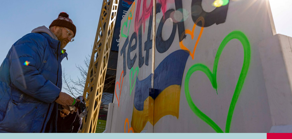 Person in a blue jacket and hat standing beside a graffiti-covered wall featuring the word 'Telford', colorful hearts, and a blue and yellow flag design under a sunny sky.