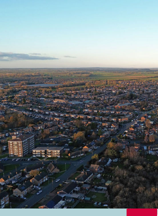Aerial view of a suburban area at sunset, showing rows of houses, streets, green spaces, and distant fields under a clear sky.