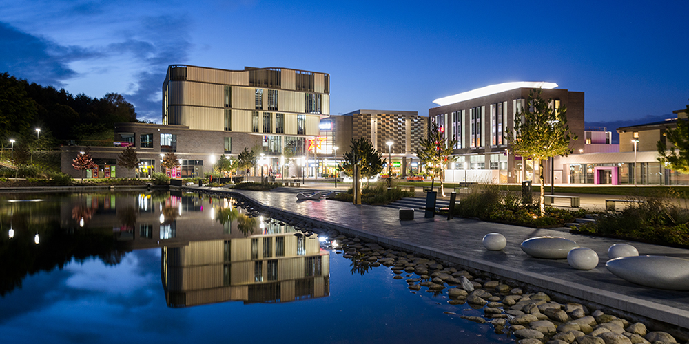 Southwater in Telford at dusk, with illuminated buildings reflected in the lake beside a landscaped waterfront walkway.