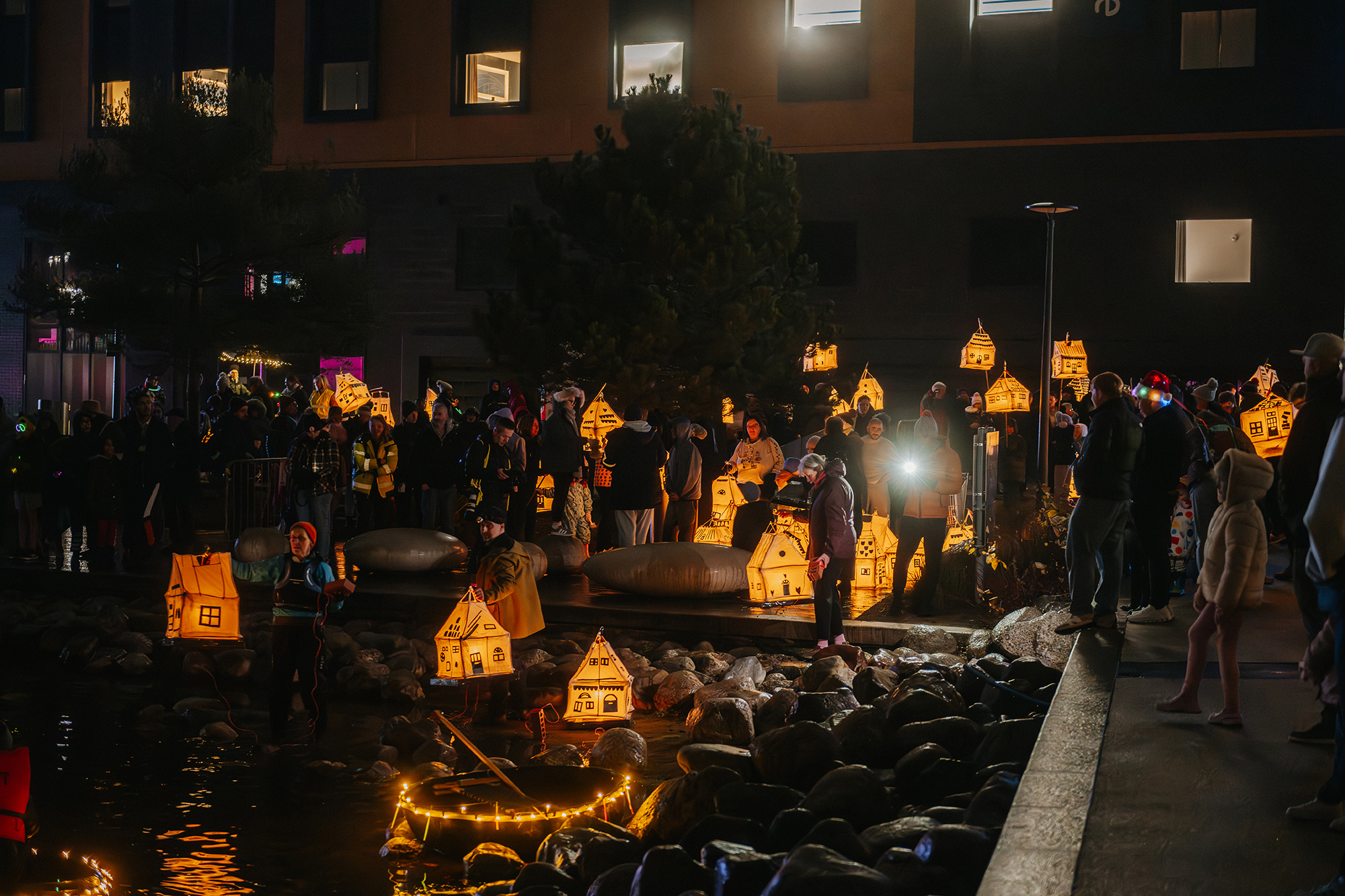 Crowds gather beside a waterfront at Telford Day 2026, carrying glowing house-shaped lanterns that illuminate the night and reflect on the water.