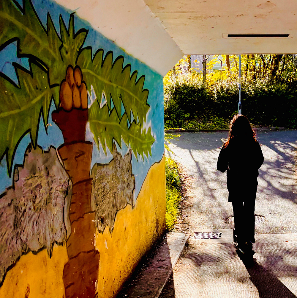 A pedestrian underpass with a colorful mural of a palm tree painted on the left wall, while a single person walks away toward a sunlit path, casting a long shadow on the ground, with trees and greenery visible beyond the tunnel.