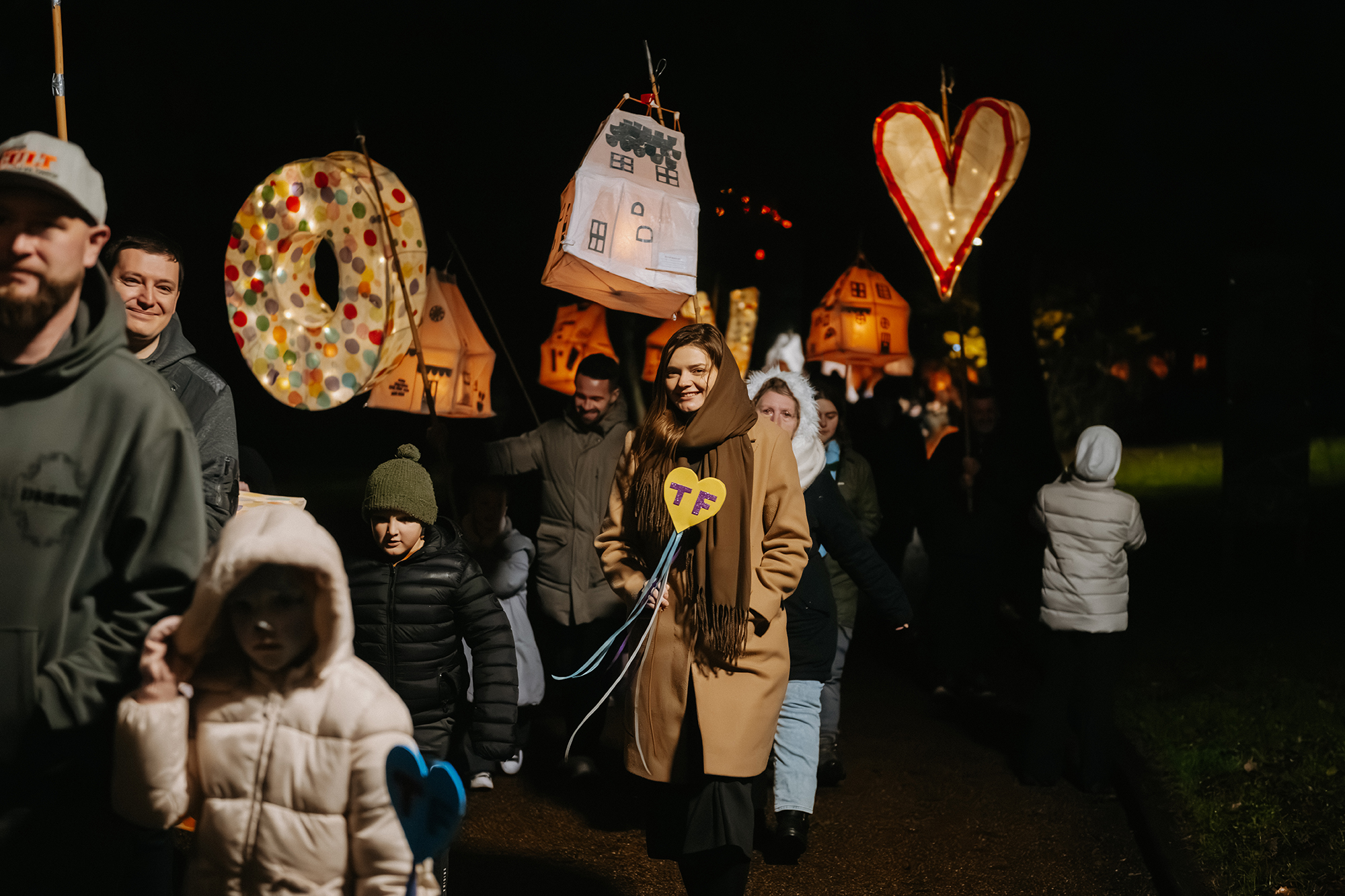 Participants in the Telford Day 2025 lantern parade carry glowing handmade lanterns shaped like houses, hearts and letters along a dark pathway.