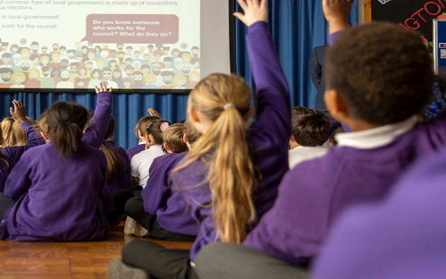A picture of school children sitting down in assembly, all putting their hands up.