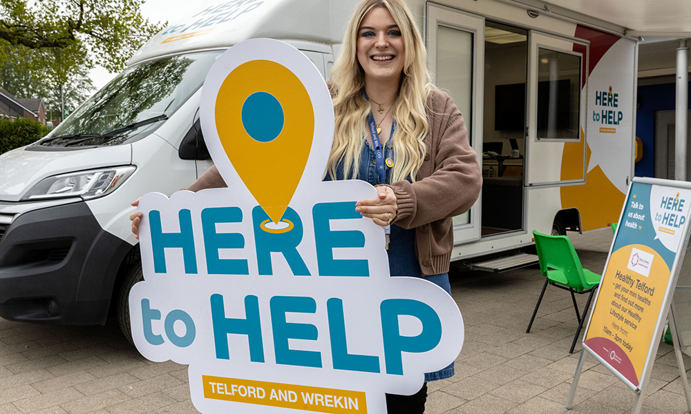 A photograph of a woman standing in front of the Here to Help bus in Telford, holding a “Here to Help” banner.