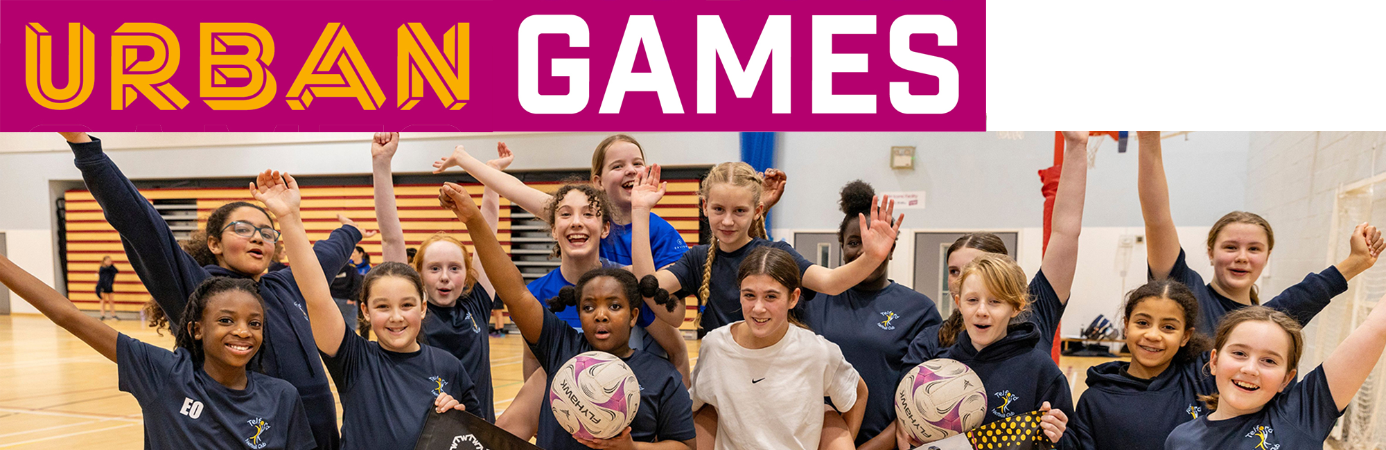 Group of children in a sports hall holding netballs and raising hands beneath a banner reading “Urban Games”.