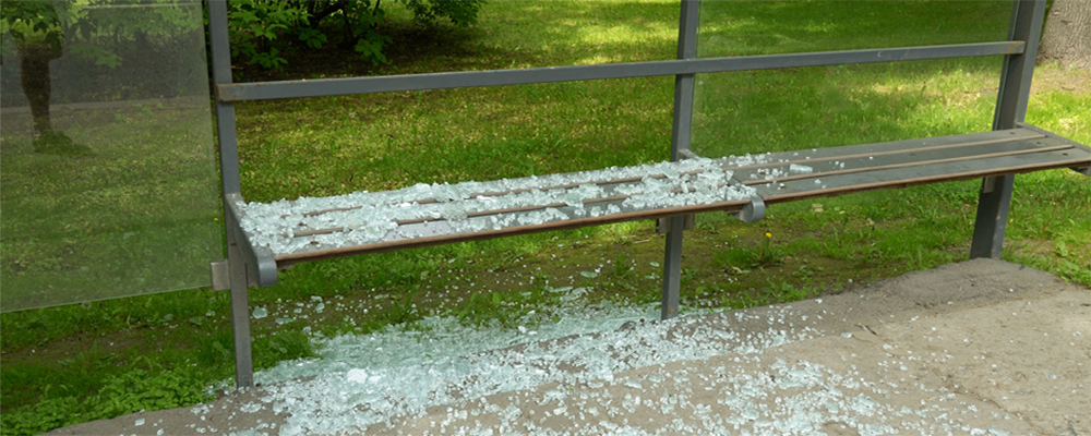 A damaged bus stop bench with broken glass pieces covering the seat and the ground around it. The bench is constructed from metal and wood and is located in a grassy, park-like setting with trees in the background.