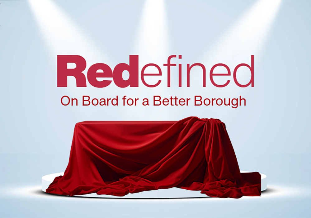 A stage setup with a red cloth covering an object at the center, highlighted by bright spotlights. Above the stage, the word 'Redefined' is displayed in bold red and black lettering, with the slogan 'On Board for a Better Borough' written beneath it.