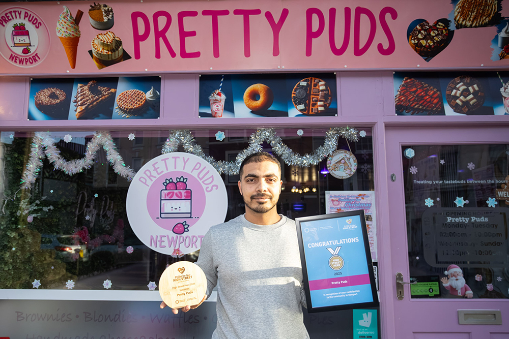 Person standing outside Pretty Puds dessert shop in Newport, holding an award plaque and certificate in front of the shop frontage.
