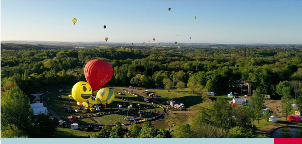 Aerial view of a hot air balloon festival in a green park surrounded by forest, featuring colorful balloons including two with yellow smiley faces and a large red one, under clear skies.