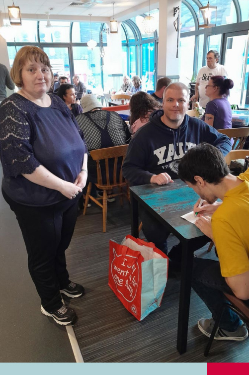 Group of people in a busy café; three individuals in the foreground, one standing and two seated at a table with a Yale hoodie and a shopping bag reading 'I went to one thing'; others are visible in the background through large windows.
